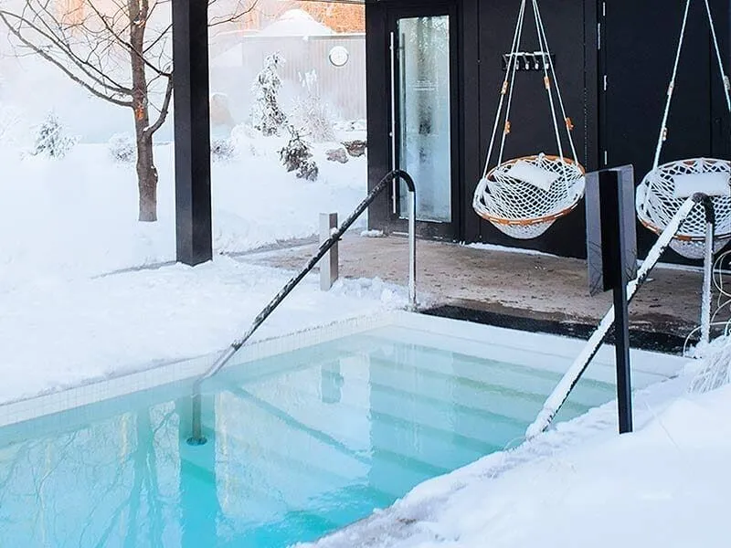 Two Cobble Mountain Summit Hammock Chairs with white pillows hang next to each other at the entrance to a cold plunge pool during winter with snow on the ground.