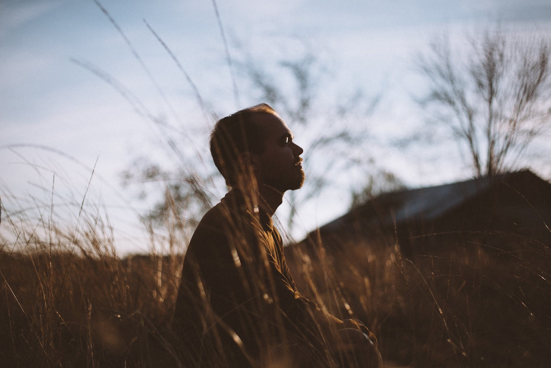 silhouette of man sitting on grass field at daytime. He appears to be taking in the sounds around him in the outdoor space.