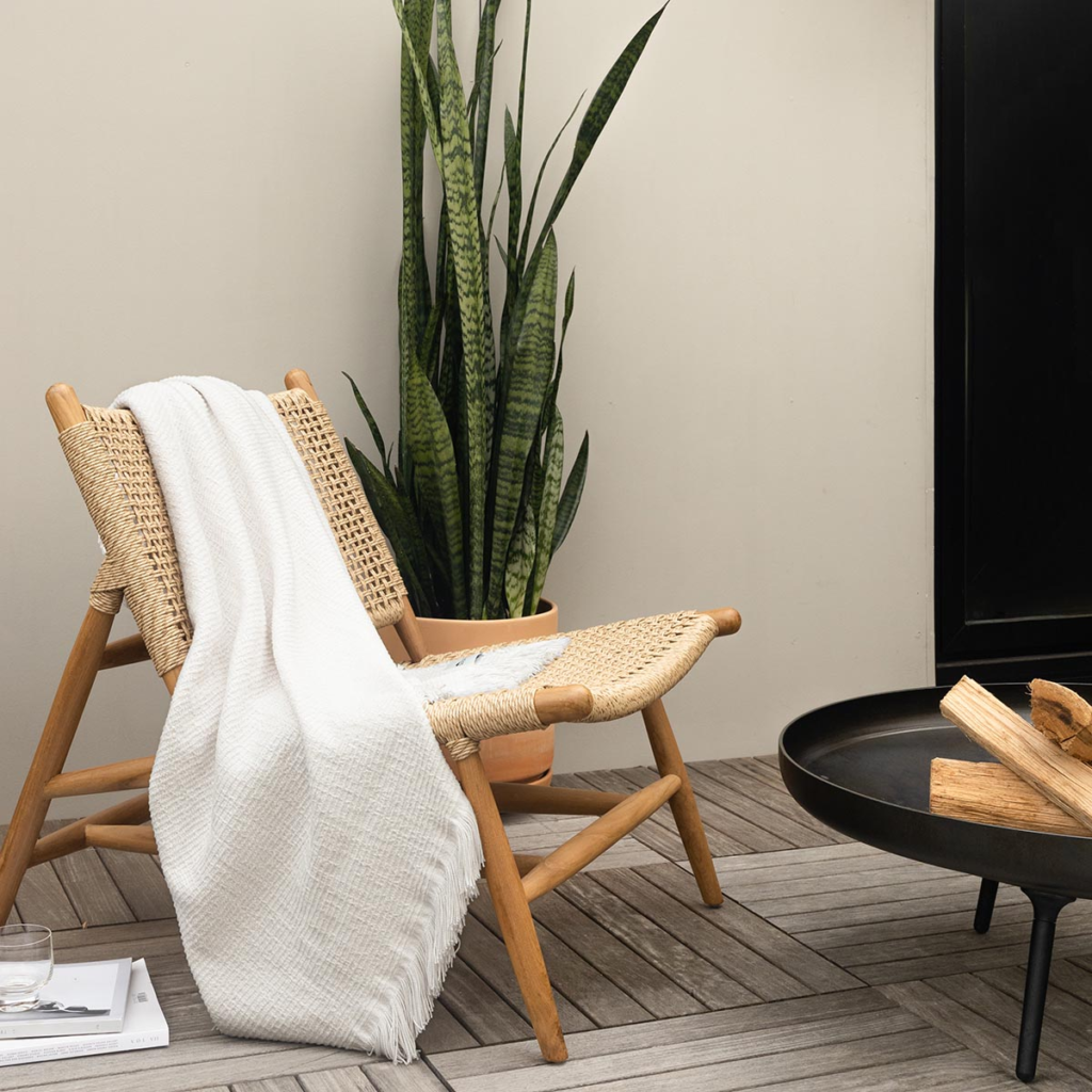Sunbrella cross hatch indoor outdoor throw blanket stylishly laid over a wooden chair in front of a firepit with logs showing.