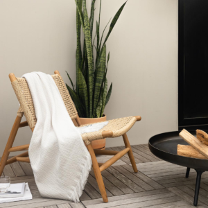 Sunbrella cross hatch indoor outdoor throw blanket stylishly laid over a wooden chair in front of a firepit with logs showing.